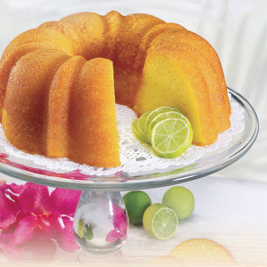 A freshly baked Key Lime Cake with a moist texture and a yellow color, decorated with a white frosting and accompanied by slices of key lime on a clear plate, with a flower and some green garnishes nearby.